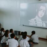 Group of school children watching anime on a projector screen indoors.